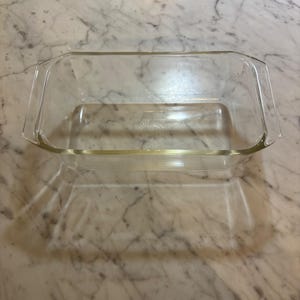 Op de afbeelding: Een rechthoekige glazen broodvorm van helder glas met geïntegreerde handgrepen aan beide korte zijden. De bodem van de vorm is voorzien van een cirkelvormig reliëflogo in het midden. De schaal is gemaakt van transparant, glad glas met een dikke rand.