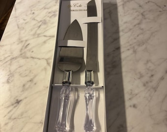 Könnte beinhalten: Ein Torten-Servierset bestehend aus einem dreieckigen Tortenheber und einem langen gezackten Messer, beide mit klaren, facettierten Acrylgriffen und Edelstahlklingen, präsentiert in einer weißen Schachtel mit der Aufschrift The Cake Set.