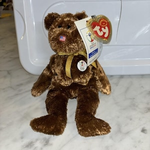 May include: A brown teddy bear with a gold ribbon and a small American flag on its nose. It has a tag indicating it's from the 2002 FIFA World Cup Korea/Japan. A collectible plush toy.