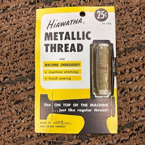 Puede incluir: Un carrete de hilo metálico dorado de Hiawatha sobre un soporte de cartón amarillo y blanco. El empaque presenta texto en negro que incluye Metallic Thread para bordado a máquina, costura a máquina y costura a mano, con un precio de 25 centavos.