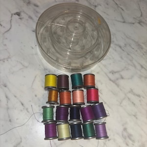 May include: A collection of seventeen small sewing thread spools in various colors including yellow, orange, red, pink, green, blue, and purple, arranged in rows below a large, round, clear plastic storage container with a lid.