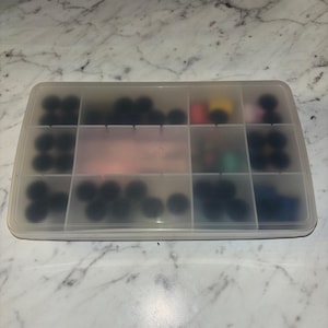 May include: A rectangular, translucent plastic storage container with multiple internal compartments. The box is filled with various small, round objects in assorted colors, including black, red, yellow, pink, green, and blue, visible through the lid.