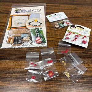 May include: A collection of craft supplies including a Blueberry Backroads nativity cross-stitch pattern, two Dress It Up holiday button sets featuring candy canes and Christmas motifs, and several small clear plastic bags containing assorted red and gold charms.