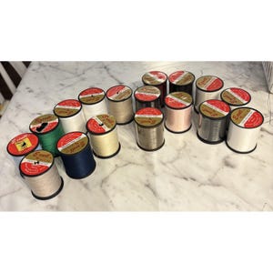 May include: A collection of seventeen spools of Excell brand 100% polyester sewing thread in various colors including white, black, grey, beige, green, and blue. Each spool features a red circular label on top with the brand name and text.