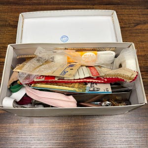 Peut inclure: Une boîte en carton blanc remplie de diverses fournitures de couture, notamment des rouleaux d'élastique blanc, des fermetures éclair roses et vertes, des emballages Dritz, une bordure rouge à accents dorés et divers petits accessoires métalliques.