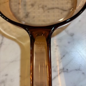 May include: A translucent amber-coloured glass saucepan featuring a long handle. The handle is embossed with the text VISION, CORNING, and FRANCE. The cookware has a smooth, rounded rim and a deep, circular basin for cooking purposes.