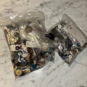 May include: Two clear plastic resealable bags filled with a large assortment of loose buttons in various sizes, shapes, and colors, including black, white, brown, blue, and green, with some buttons featuring four holes and others featuring two holes.