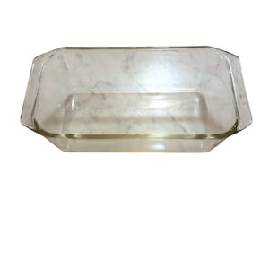 May include: A clear, rectangular glass loaf pan with beveled corners and integrated handles on the short ends. The base of the pan features a circular embossed logo in the centre, and the glass has a smooth, transparent finish throughout.