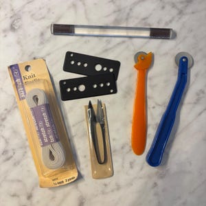 May include: A sewing kit containing a package of 1/4 inch Knit Elastic, two black rectangular metal plates with circular cutouts, a pair of small metal snips, a clear plastic rod, and two tracing wheels with orange and blue plastic handles.