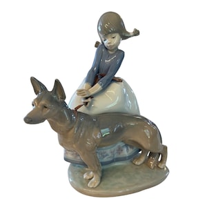 May include: A porcelain figurine depicting a young girl in a blue dress with a white skirt, holding a leash attached to a gray dog. The figurine is on a light green base and has a detailed design.