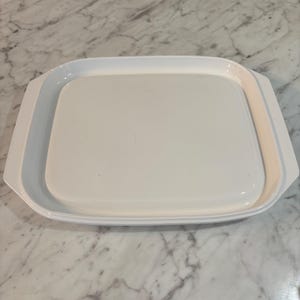 Könnte beinhalten: Ein weißes, quadratisches Serviertablett mit abgerundeten Ecken und integrierten Seitengriffen. Das Tablett verfügt über eine glatte, glänzende Oberfläche und einen leicht vertieften Mittelbereich, was ein klares und funktionales Design ergibt.