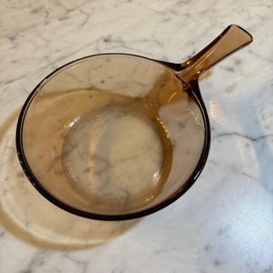 Puede incluir: Un cuenco redondo de cristal transparente de color ámbar con un asa integrada que se extiende desde el lateral. El cuenco tiene una superficie lisa y pulida y una parte superior ancha y abierta, mostrando un tono marrón claro uniforme.