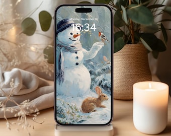 Pode incluir: Um smartphone exibe uma cena de inverno com um boneco de neve, um pássaro e um coelho. A tela mostra a data e hora: segunda-feira, 18 de dezembro, 16:34. O telefone está em um suporte branco, com uma vela acesa e uma planta em um vaso tecido por perto.