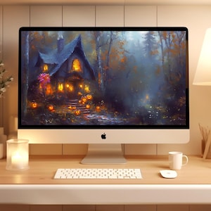 May include: A silver desktop computer monitor displaying a digital illustration of a dark, atmospheric cottage in a forest at night, surrounded by glowing orange pumpkins, set on a wooden desk with a white keyboard, mouse, and a lamp.