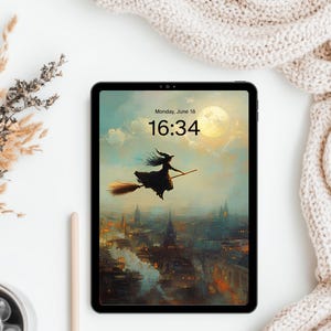 May include: A black tablet displaying a digital illustration of a witch flying on a broomstick over a city at night with a full moon. The screen shows the text Monday, June 18 and the time 16:34. The device has a thin black frame and rounded corners.