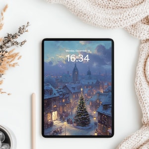May include: A black tablet displays a winter scene with a Christmas tree and buildings. The screen shows the time 16:34 and the date, Monday, December 18. A cup of coffee, a pen, and dried plants are on a white surface.
