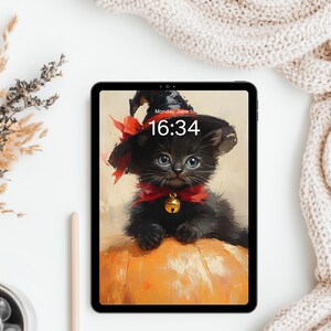 May include: A black tablet displaying a digital illustration of a fluffy black kitten wearing a witch hat with a red ribbon and a gold bell, perched on a large orange pumpkin. The screen shows the time 16:34 and the date Monday, June 18.