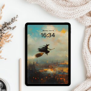 May include: A black tablet displaying a digital illustration of a witch flying on a broomstick over a city at sunset. The screen shows the text Monday, June 18 and 16:34. The tablet is positioned on a white surface with dried plants and fabric.