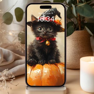 May include: A smartphone displaying a digital wallpaper of a fluffy black kitten wearing a witch hat and a red bow with a bell, sitting on a large orange pumpkin. The screen shows the time 16:34 and the date Monday, June 18 in white text.
