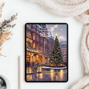 Può includere: Un tablet digitale mostra una scena invernale con un albero di Natale decorato e un edificio illuminato. Lo schermo mostra l'ora: 16:34. Una tazza di caffè, una penna e piante secche sono su una superficie bianca accanto a una coperta di maglia color crema.