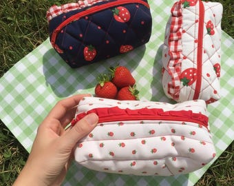 May include: Three quilted cosmetic bags with strawberry patterns. One is navy blue with red strawberries, another is white with red strawberries, and the third is white with red strawberries and a red ruffle. Fresh strawberries are next to the bags.