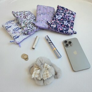 Puede incluir: Un surtido de bolsas de tela acolchadas con estampados florales en lavanda, blanco y azul marino. También se ve un iPhone plateado, un accesorio con forma de conejo gris esponjoso con un lazo, monedas y dos frascos de perfume plateados.