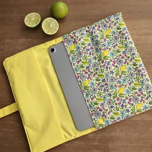 Può includere: Una custodia gialla per iPad con un motivo floreale di limoni, fiori rosa e blu. All'interno è visibile un tablet grigio. Due metà di lime e un lime intero sono su una superficie di legno.