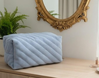 May include: A light blue, rectangular cosmetic bag with a quilted, diagonal pattern. The bag is made of a soft, corduroy-like material. The bag is sitting on a wooden surface, with a gold-framed mirror in the background.