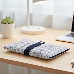 Puede incluir: Una funda acolchada para portátil con un estampado floral azul y blanco. La funda tiene una correa azul marino con un cierre de botón blanco. Está sobre un escritorio de madera, con una lámpara de escritorio y un portátil al fondo.