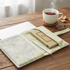 Pode incluir: Um livro intitulado "Gucci" numa capa de livro xadrez verde e branco, uma caneta stylus branca e uma chávena de chá branca numa mesa de madeira. A capa do livro está aberta, revelando um interior estampado. Uma chávena de chá fumega por perto.