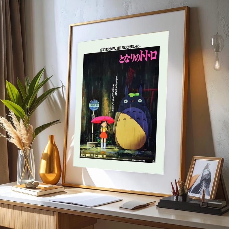 May include: Framed poster featuring a scene from "My Neighbor Totoro." The artwork depicts a young girl with a red umbrella, standing near a bus stop with a large, furry creature. Japanese text is visible at the top.
