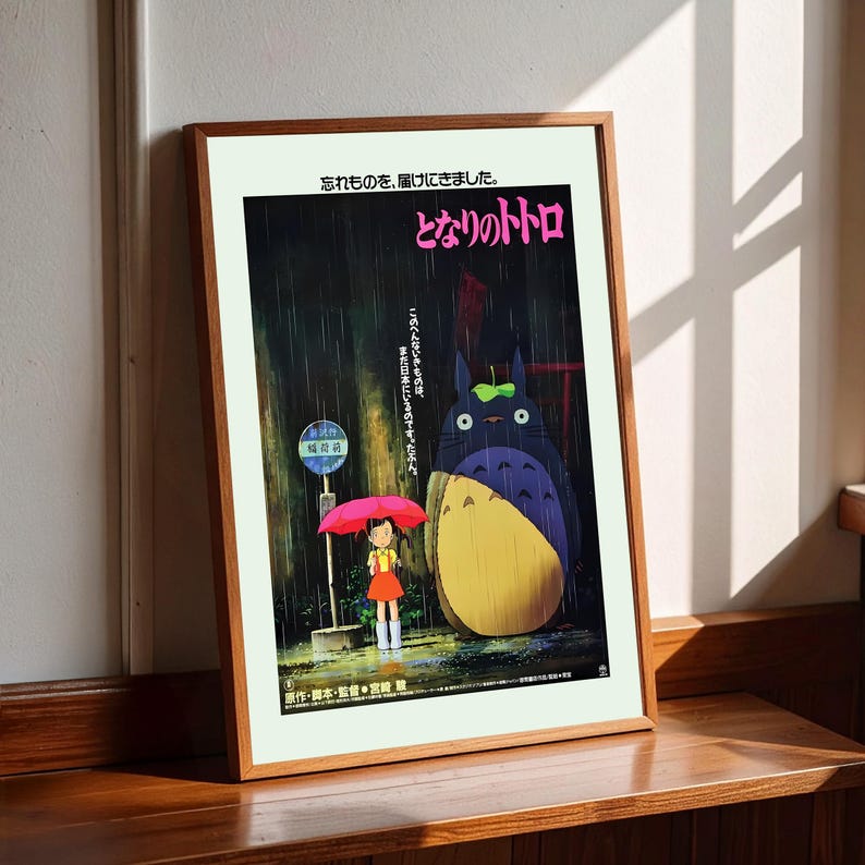 May include: Framed poster featuring the Japanese animated film "My Neighbor Totoro." The artwork depicts Totoro, a large forest spirit, standing next to a young girl with an umbrella in the rain. Japanese text is visible.