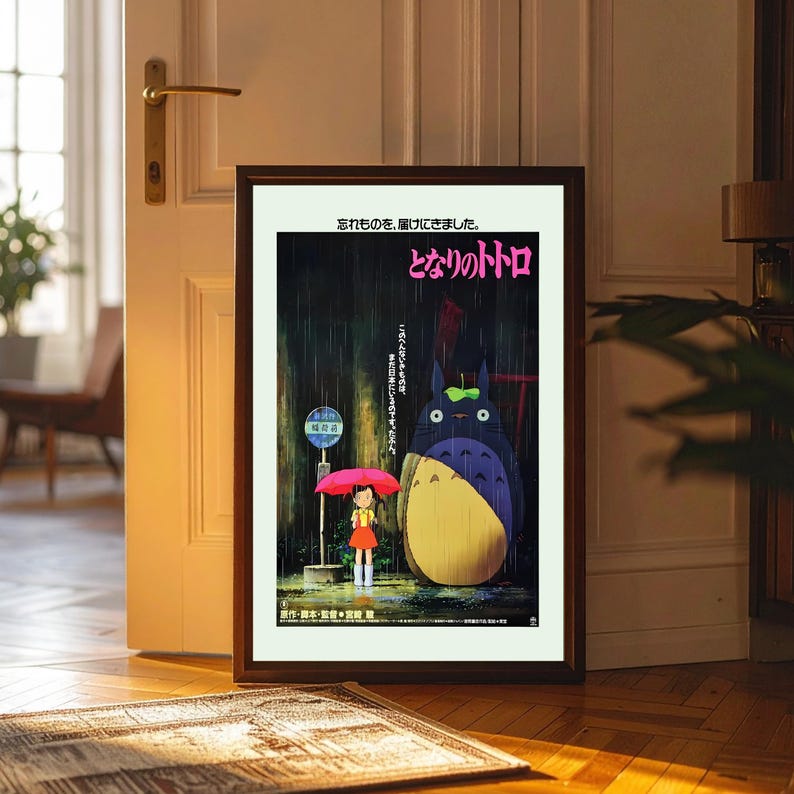 May include: Framed poster featuring the animated film "My Neighbor Totoro." The artwork depicts a young girl with a red umbrella, standing in the rain next to a large, furry creature. Japanese text is visible at the top and bottom of the poster.
