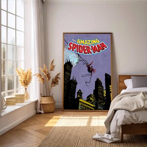 May include: A framed poster featuring the comic book cover of "The Amazing Spider-Man." The artwork depicts Spider-Man swinging above a cityscape with a purple background. The poster is displayed in a well-lit room with a window and bed.