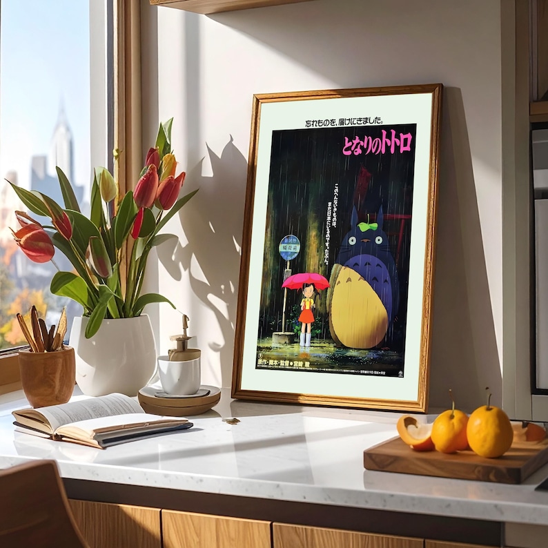May include: Framed poster featuring a scene from the animated film "My Neighbor Totoro." The artwork depicts Totoro, a large forest spirit, and a young girl under an umbrella in the rain. The poster is in a gold frame, displayed on a countertop.