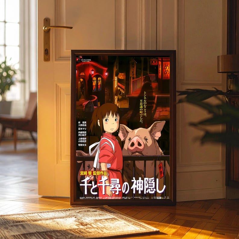 May include: Framed poster featuring artwork from the animated film "Spirited Away." The poster shows a young girl in a red outfit with a pig, set against a backdrop of a Japanese town. The title is in Japanese.
