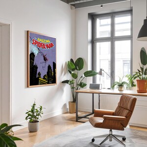 May include: A framed comic book art print of The Amazing Spider-Man. The artwork features Spider-Man swinging over a city skyline with a purple background. The frame is a light brown color and is hanging on a white wall.