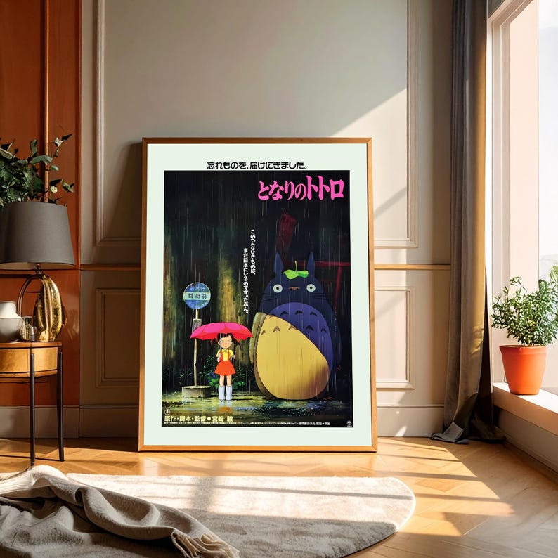 May include: Framed poster featuring the animated film "My Neighbor Totoro." The artwork depicts a young girl with a red umbrella standing at a bus stop in the rain, with Totoro, a large forest spirit, nearby. Japanese text is visible.