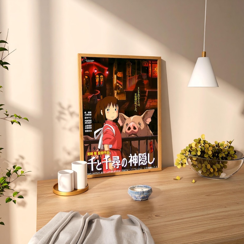 May include: A framed poster featuring a scene from the animated film "Spirited Away." The artwork depicts a young girl in a red outfit, a large pig, and Japanese text. The poster is in a wooden frame, displayed on a wooden table.