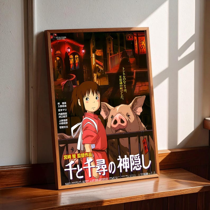 May include: Framed poster featuring artwork from the animated film "Spirited Away." The poster shows a young girl in a red outfit, a large pig, and Japanese text. The poster is in a wooden frame and is displayed on a wooden surface.