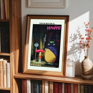 May include: Framed poster featuring the animated film "My Neighbor Totoro." The artwork depicts a young girl with a red umbrella standing near a bus stop, with Totoro in the rain. Japanese text is visible at the top and right of the poster.