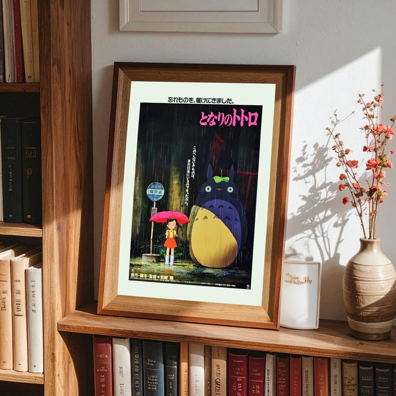May include: Framed poster featuring the animated film "My Neighbor Totoro." The artwork depicts a young girl with a red umbrella standing near a bus stop, with Totoro in the rain. Japanese text is visible at the top and right of the poster.