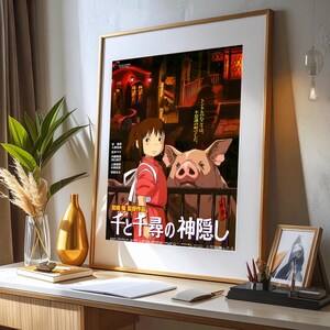 May include: Framed poster featuring artwork from the animated film "Spirited Away." The image shows a young girl in a red outfit, a large pig, and Japanese text. The poster is in a gold frame and is displayed on a white desk.