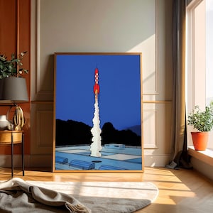 Puede incluir: Una impresión artística enmarcada representa un cohete rojo y blanco despegando hacia un cielo azul oscuro. El cohete emite una columna de humo blanco mientras asciende. La obra de arte presenta un estilo retro, tipo caricatura, con una silueta negra de montañas.