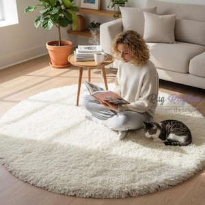 Puede incluir: Una alfombra redonda y crema con pelo largo sobre un suelo de madera. Una persona está sentada en la alfombra, leyendo un libro. Un gato está tumbado en la alfombra. Una pequeña mesa de madera con una taza y libros está cerca. Una planta en maceta está en el fondo.
