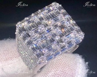 May include: A large, rectangular silver ring encrusted with numerous rectangular-cut, clear gemstones. The ring's surface is densely packed with the stones, creating a sparkling, textured appearance. The ring is set against a dark background.