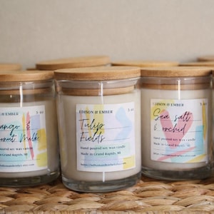 Puede incluir: Una colección de velas en tarros de vidrio con tapas de madera. Cada vela tiene una etiqueta con un nombre de aroma diferente, como "Mango & Coconut Milk" y "Tulip Fields". Las etiquetas presentan diseños abstractos coloridos. Las velas son de 142 gramos.