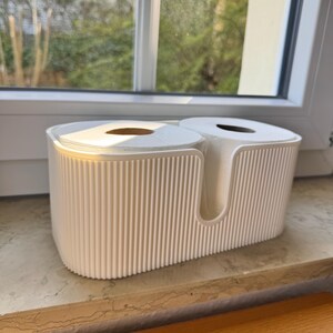 May include: A white, oval-shaped toilet paper holder with a ribbed, vertical texture on the exterior. The container features a U-shaped cutout on the front and is designed to hold two standard rolls of white toilet paper side by side.
