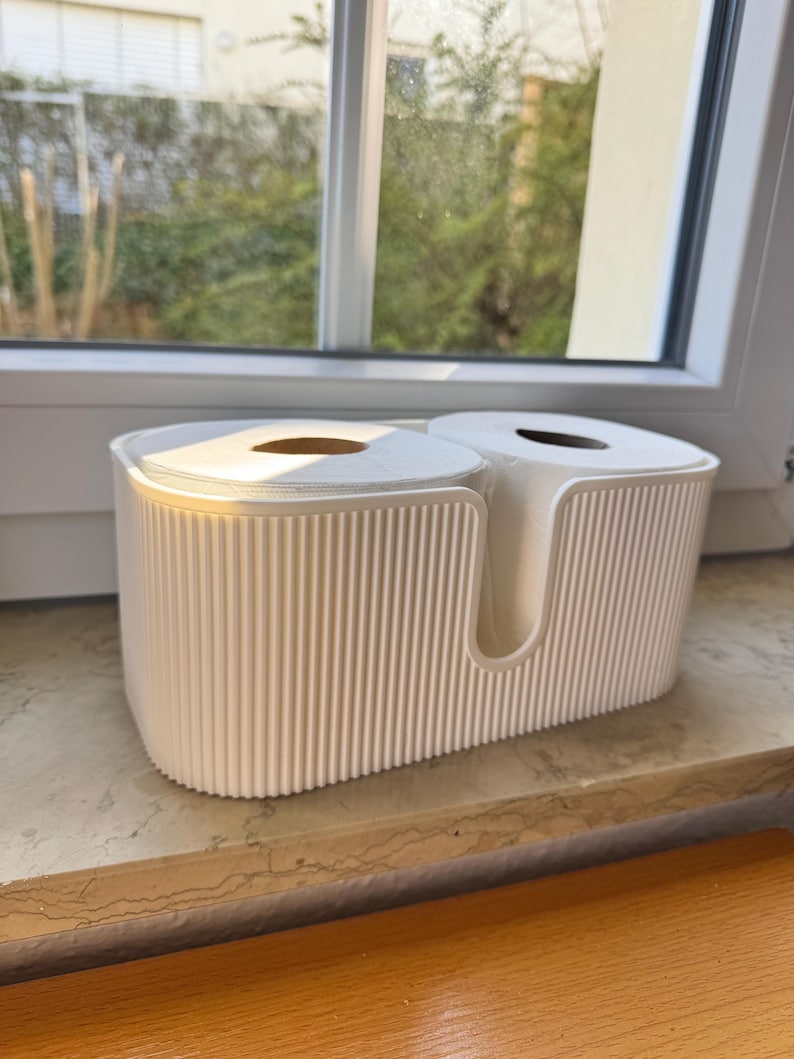 May include: A white, oval-shaped toilet paper holder with a ribbed, vertical texture on the exterior. The container features a U-shaped cutout on the front and is designed to hold two standard rolls of white toilet paper side by side.