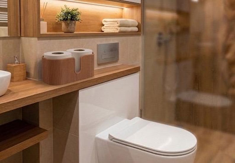 May include: A brown, ribbed, oval-shaped toilet paper holder containing two rolls of white toilet paper. The holder features a U-shaped cutout on the front and is positioned on a wooden bathroom counter next to a white toilet and a glass shower.